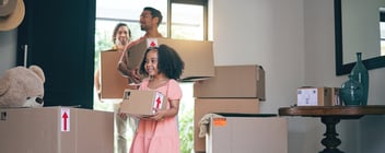 Two adults and a child walk into a home while carrying brown moving boxes